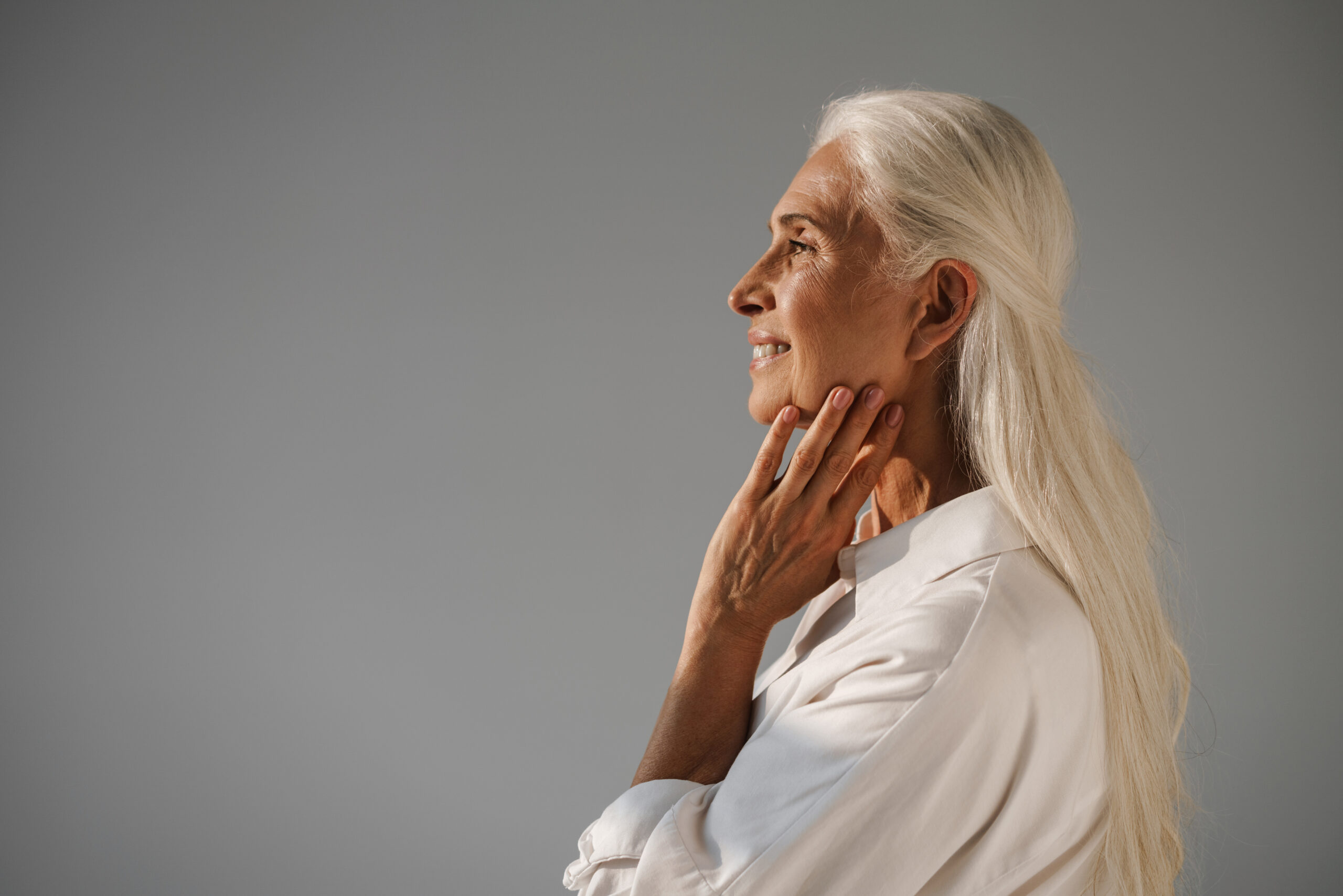 Profile of smiling senior woman with long white hair, touching her neck, looking thoughtful against gray background.