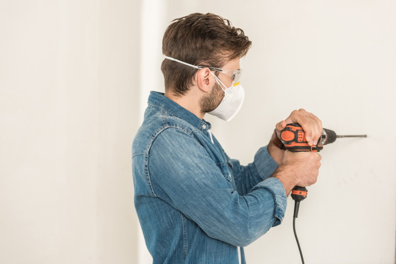 young man in protective workwear using electric drill at wall during house repair